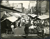 View of the stalls and market goers around the perimeter of Lexington Market in Baltimore, Maryland. Vendors in the foreground can be seen selling a variety of fruits and vegetables.