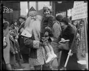 Santa Claus holding a young girl on a crowded sidewalk. Part of an American Rescue Worker sign and donation bucket are on the right. Likely Howard Street or Lexington Street in Baltimore, Maryland.