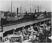 View of the Piedmont Air Line railroad dock at Light Street in Baltimore, Maryland. The sign above the docks reads "Pier 2 / Piedmont Air Line / via York River / Richmond & Danville Rail Road." People stand in front of the building alongside horses, wagons, and barrels. A sign that runs underneath the roof…
