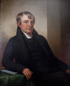 Three-quarter length seated portrait of Edward Johnson (1767-1829). He is portrayed as a man with short wavy gray hair, with one lock brushed over forehead. He wears black coat, pants, and vest over a white shirt. He props his proper right elbow and arm on a table covered with a green cloth. Johnson was a…