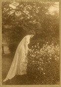 Outdoor portrait of Sue Collins, a friend of photographer Emily Spencer Hayden. Collins stands outside in a long, white dress and bends away from the camera to pick a flower.