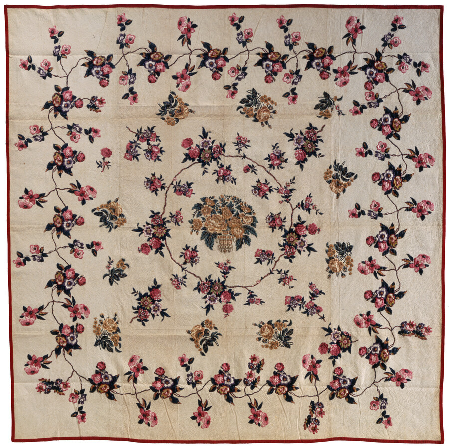 Quilt decorated with floral chintz applique on a plain white cotton background with a thin red border. A central vase of flowers motif is surrounded by three rows of floral vines.