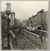 View of the 600 block of Baltimore Street, looking east, in the downtown area of Baltimore City, Maryland. Trolley tracks line the street and a trolley is visible in the distance. There are a number of shops that tightly pack both sides of the street. On the left side of the photo, the sign "National…