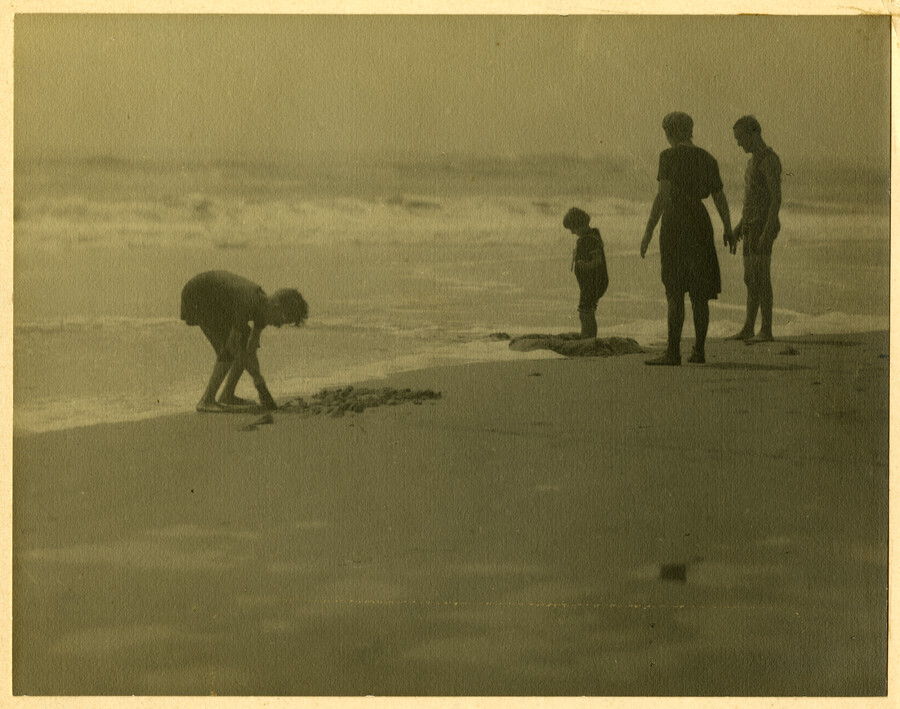 On the shore, a group of beachgoers stand near the surf. The group is presumed to be the family of Baltimore, Maryland, photographer Emily Spencer Hayden. Verso transcription: Along The Shore - Hayden