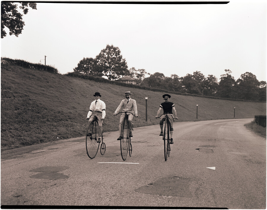 Richard Inglehart, Donald Lang, and Charles Logue riding high wheelers (also known as penny-farthings), re-enacting the "Gay '90s" at Clifton Lake in Baltimore, Maryland.