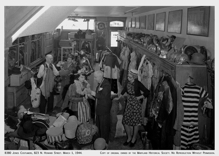 Interior view of A. T. Jones and Sons costume company, once located on 823 North Howard Street in Baltimore, Maryland. Customers can be seen shopping and trying on costumes. While at this location, the shop had 17 different rooms full of costume materials. The company was founded in 1868 and had one of the largest…