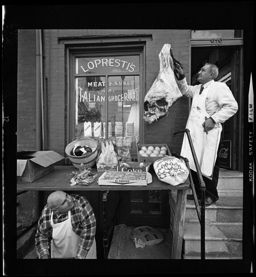 Aproned grocers display their goods outside Lopresti's Meat Market and Italian Grocery in Little Italy, Baltimore, Maryland. The shopkeeper on the right hangs a large leg of meat, a second shopkeeper stands in the cellar stairwell. Resting on the stoop stairs is a makeshift table displaying eggs, Coca-Cola, and produce.